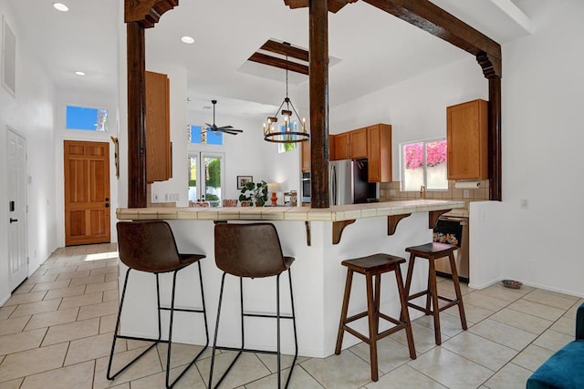 kitchen with tile countertops, a kitchen bar, stainless steel refrigerator, light tile patterned floors, and ceiling fan with notable chandelier