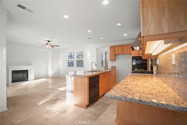 kitchen featuring wall chimney exhaust hood, sink, light stone counters, a center island with sink, and black appliances