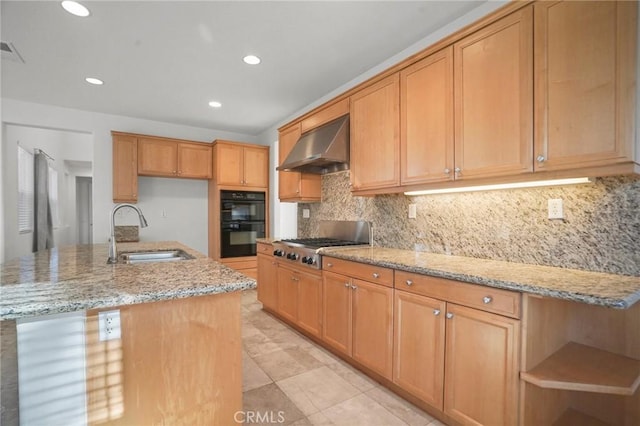 kitchen with sink, black double oven, a kitchen island with sink, light stone counters, and wall chimney exhaust hood