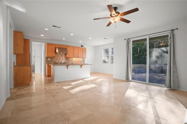 kitchen with a breakfast bar, ceiling fan, wall chimney range hood, light stone countertops, and backsplash