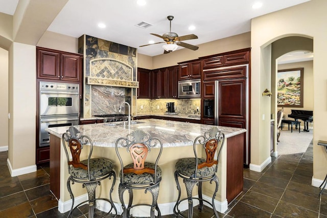 kitchen with tasteful backsplash, light stone countertops, stainless steel appliances, and a kitchen island with sink