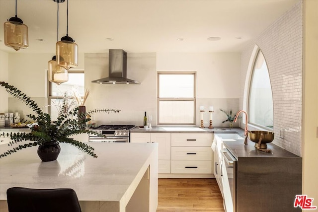 kitchen featuring decorative light fixtures, white cabinetry, wall chimney range hood, stainless steel gas stove, and light hardwood / wood-style flooring