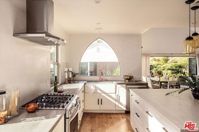 kitchen with high end stove, hanging light fixtures, wall chimney exhaust hood, white cabinets, and sink