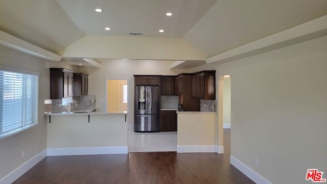 kitchen with kitchen peninsula, dark brown cabinets, vaulted ceiling, stainless steel fridge, and tasteful backsplash