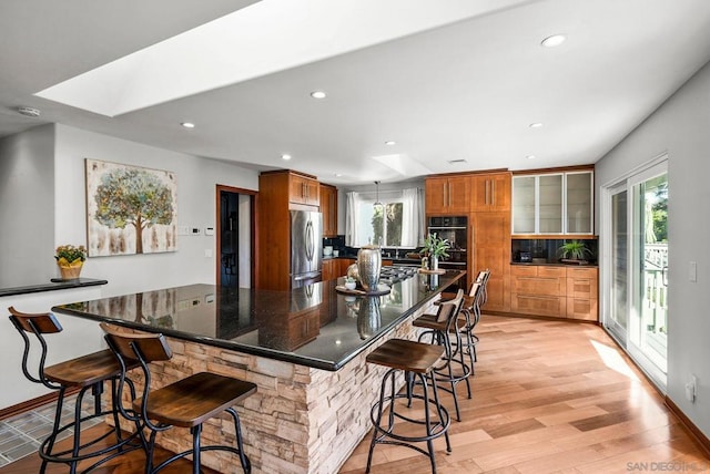 kitchen featuring a skylight, dark stone countertops, stainless steel fridge, light hardwood / wood-style flooring, and a breakfast bar area