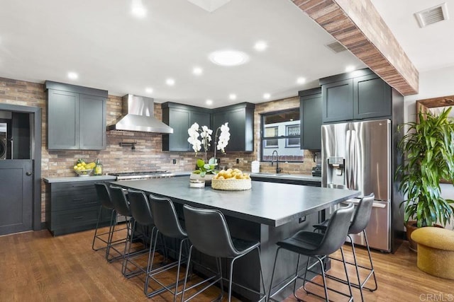 kitchen with a kitchen island, wood-type flooring, a kitchen bar, stainless steel appliances, and wall chimney exhaust hood