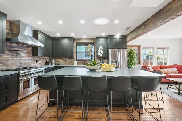 kitchen featuring wall chimney range hood, appliances with stainless steel finishes, a center island, a kitchen bar, and dark hardwood / wood-style flooring