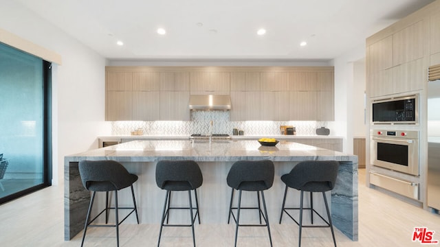 kitchen with light stone countertops, exhaust hood, stainless steel oven, a center island with sink, and built in microwave