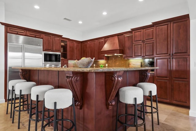 kitchen with light stone countertops, a large island with sink, built in appliances, custom range hood, and a breakfast bar area