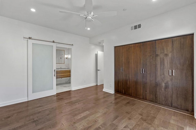 unfurnished bedroom with ensuite bath, hardwood / wood-style flooring, a closet, ceiling fan, and a barn door