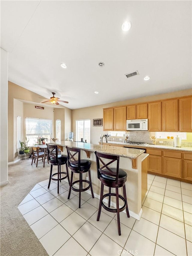 kitchen with a center island with sink, ceiling fan, a kitchen breakfast bar, light colored carpet, and light stone counters