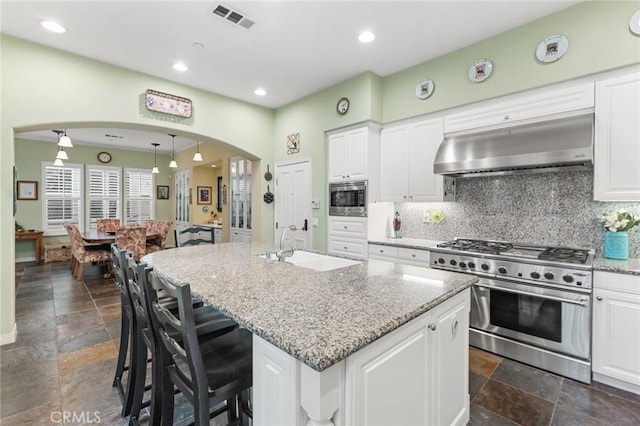 kitchen featuring arched walkways, a kitchen island with sink, stainless steel appliances, exhaust hood, and white cabinets