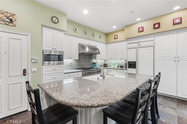 kitchen with a breakfast bar, light stone counters, a center island with sink, stainless steel microwave, and range hood