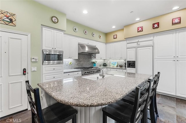 kitchen featuring a breakfast bar, range hood, a center island with sink, stainless steel microwave, and light stone countertops