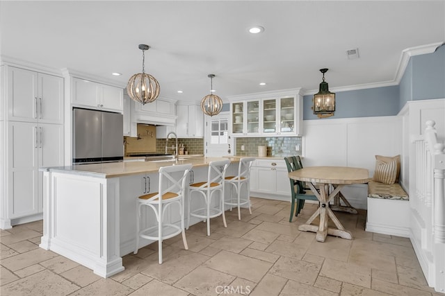 kitchen with stainless steel fridge, a breakfast bar area, an island with sink, white cabinets, and decorative light fixtures