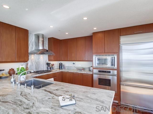 kitchen featuring wall chimney range hood, kitchen peninsula, sink, built in appliances, and light stone countertops