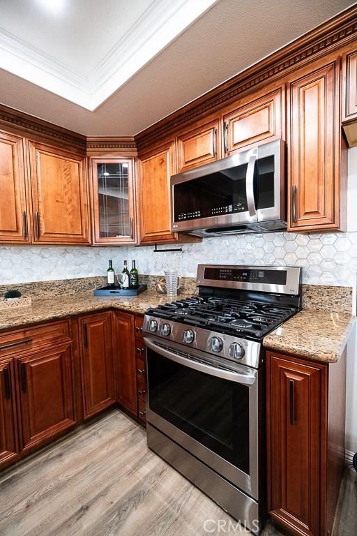 kitchen featuring light hardwood / wood-style flooring, appliances with stainless steel finishes, a tray ceiling, ornamental molding, and light stone counters