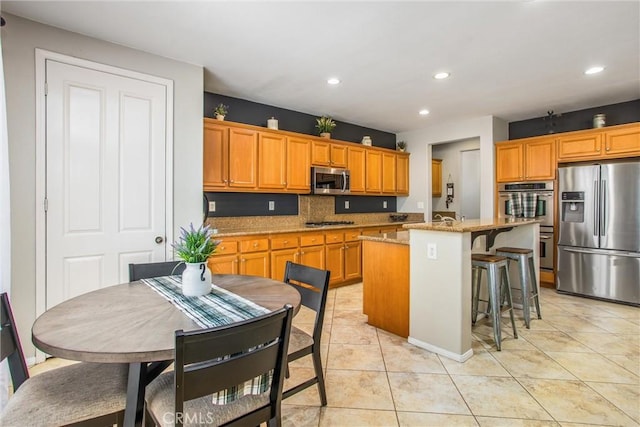 kitchen featuring light tile patterned floors, appliances with stainless steel finishes, decorative backsplash, a kitchen island with sink, and light stone countertops