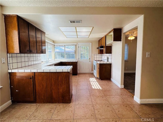 kitchen featuring tile countertops, kitchen peninsula, tasteful backsplash, light tile patterned flooring, and white gas range oven