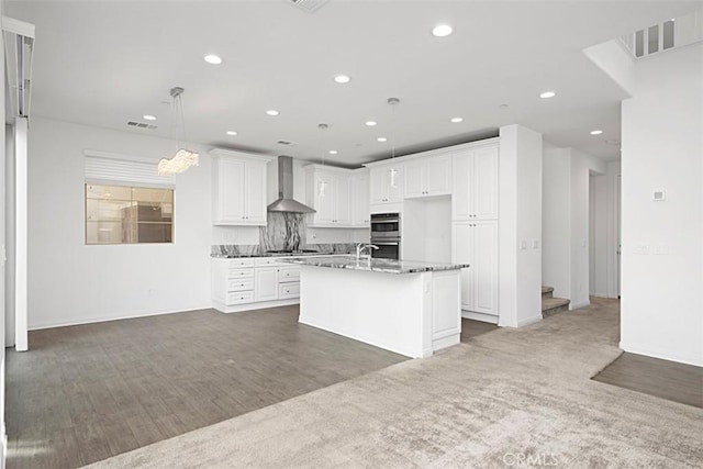 kitchen with white cabinets, wall chimney range hood, hanging light fixtures, a kitchen island with sink, and stone counters