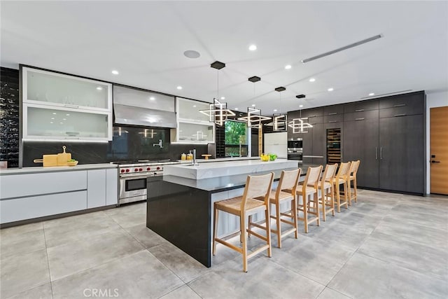 kitchen featuring a center island with sink, range with two ovens, decorative light fixtures, wall chimney range hood, and a breakfast bar