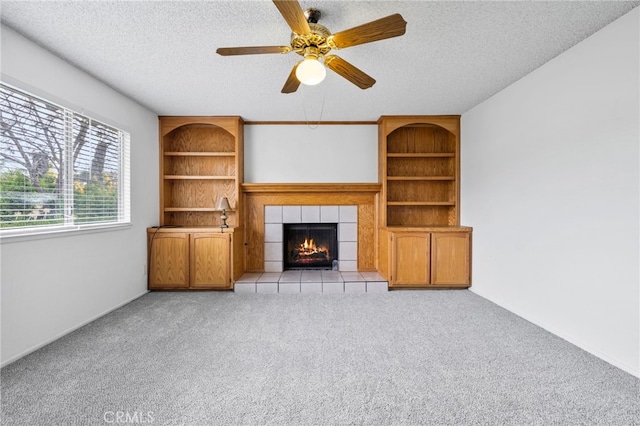 unfurnished living room with ceiling fan, light colored carpet, a tiled fireplace, built in features, and a textured ceiling