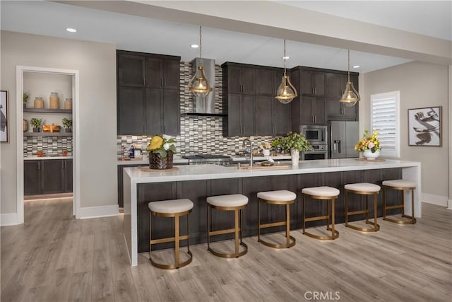 bar with pendant lighting, wall chimney range hood, stainless steel appliances, light wood-type flooring, and dark brown cabinets