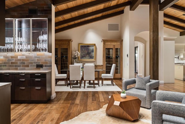 bar with light hardwood / wood-style floors, tasteful backsplash, dark brown cabinets, stainless steel counters, and high vaulted ceiling