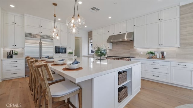 kitchen with white cabinets, decorative light fixtures, built in appliances, a kitchen island with sink, and light hardwood / wood-style flooring