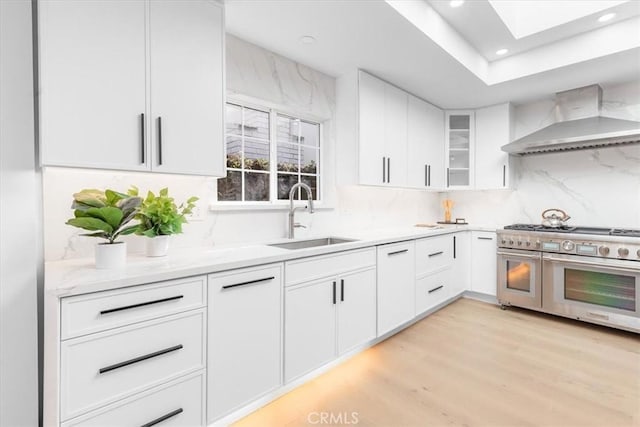 kitchen featuring sink, wall chimney range hood, white cabinets, and range with two ovens