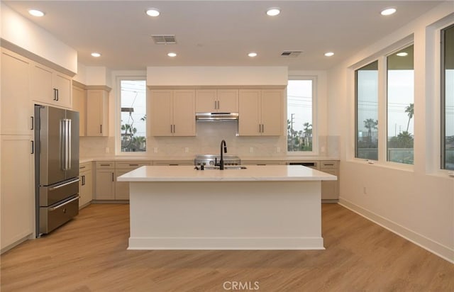 kitchen with tasteful backsplash, high end refrigerator, sink, light wood-type flooring, and an island with sink