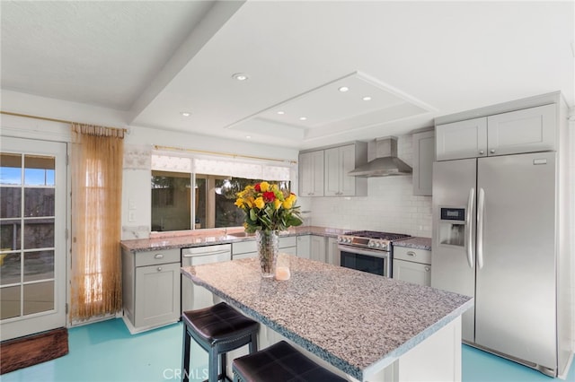 kitchen featuring a kitchen bar, appliances with stainless steel finishes, a raised ceiling, wall chimney exhaust hood, and a center island