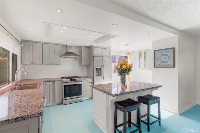 kitchen with wall chimney range hood, a raised ceiling, sink, appliances with stainless steel finishes, and light stone counters