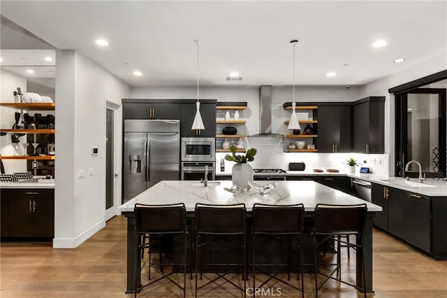 kitchen featuring sink, built in appliances, decorative light fixtures, an island with sink, and wall chimney range hood