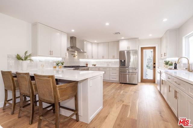 kitchen with a breakfast bar area, stainless steel appliances, white cabinetry, and wall chimney range hood