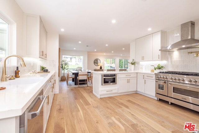 kitchen featuring wall chimney range hood, kitchen peninsula, sink, appliances with stainless steel finishes, and white cabinets