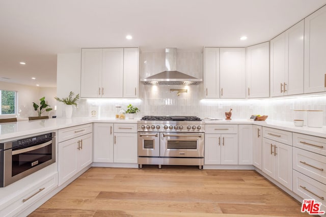 kitchen with white cabinetry, appliances with stainless steel finishes, decorative backsplash, light wood-type flooring, and wall chimney range hood