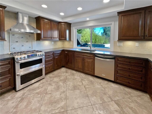 kitchen featuring wall chimney range hood, sink, backsplash, stainless steel appliances, and a tray ceiling