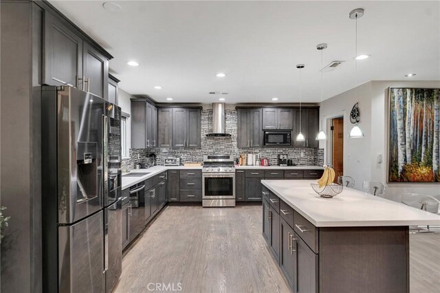 kitchen with wall chimney range hood, stainless steel appliances, decorative backsplash, hanging light fixtures, and light wood-type flooring