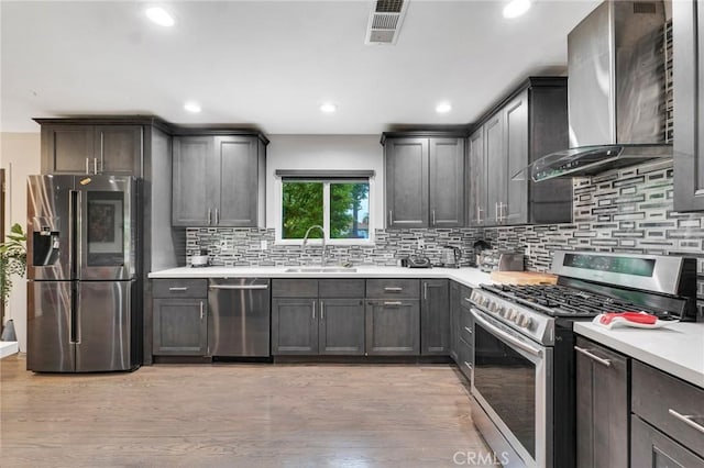 kitchen with appliances with stainless steel finishes, wall chimney range hood, decorative backsplash, sink, and light hardwood / wood-style flooring