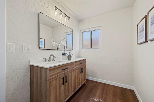 full bathroom featuring wood finished floors, baseboards, double vanity, a sink, and a textured wall