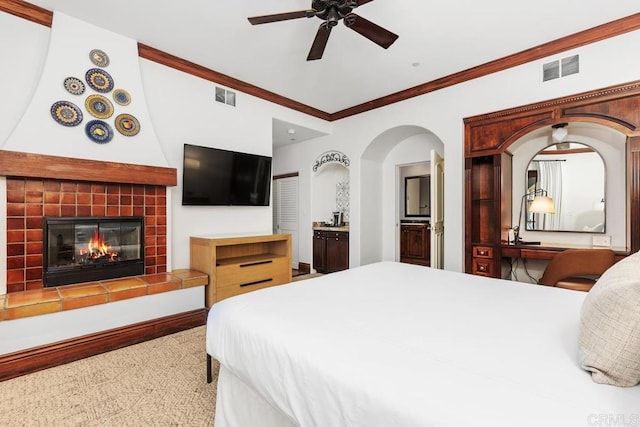 bedroom featuring a tiled fireplace, crown molding, ceiling fan, and ensuite bath