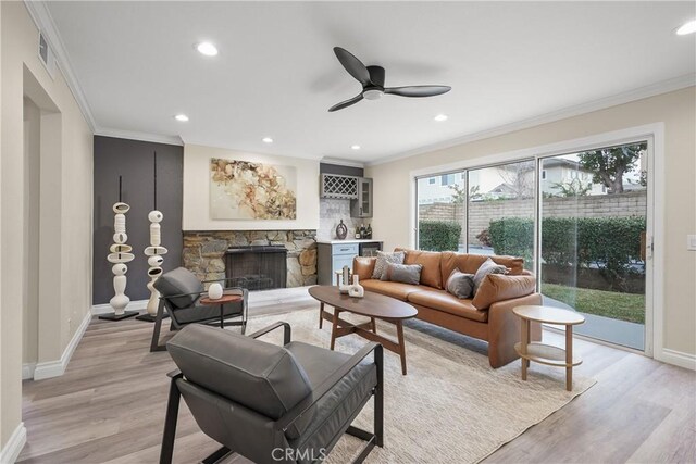 living room with ceiling fan, crown molding, light wood-type flooring, a stone fireplace, and bar