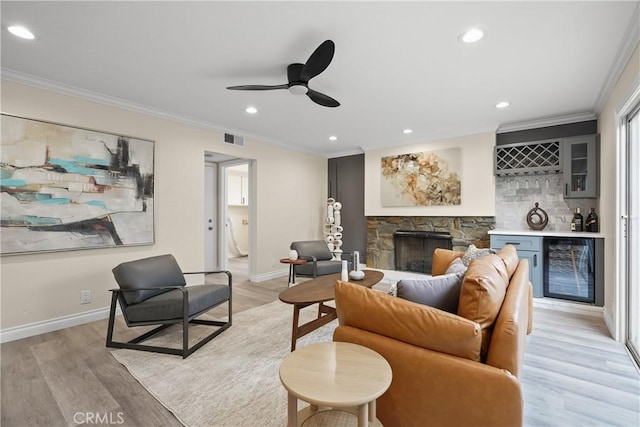 living room with beverage cooler, a stone fireplace, light wood-type flooring, bar area, and crown molding