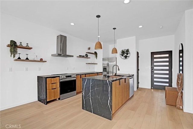 kitchen with wall chimney range hood, light hardwood / wood-style floors, a center island with sink, sink, and stainless steel appliances