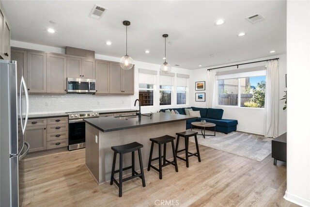 kitchen with stainless steel appliances, sink, hanging light fixtures, light wood-type flooring, and a center island with sink