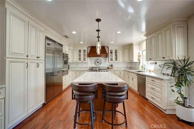 kitchen featuring a kitchen island, custom exhaust hood, hanging light fixtures, light stone countertops, and stainless steel appliances