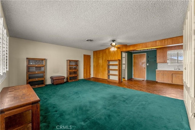 unfurnished living room featuring a textured ceiling, dark colored carpet, sink, wood walls, and ceiling fan