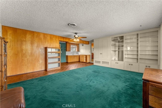 unfurnished living room with wood walls, ceiling fan, dark colored carpet, built in features, and a textured ceiling