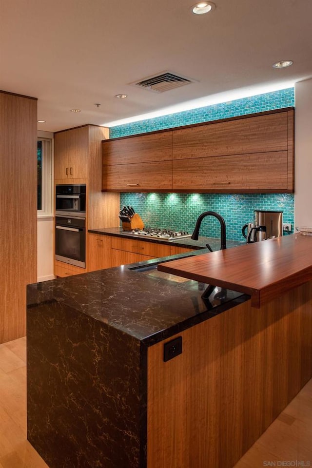 kitchen with black double oven, light hardwood / wood-style floors, backsplash, kitchen peninsula, and stainless steel gas cooktop